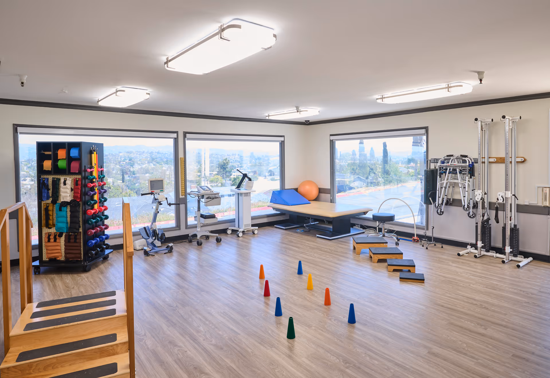 A spacious physical therapy or rehabilitation room with large windows showing an outdoor view. The room contains exercise equipment including a stationary bike, weights, resistance bands, therapy steps, cones for agility drills, a therapy table, and walkers. The floor is wood, and the room is well-lit with ceiling lights.