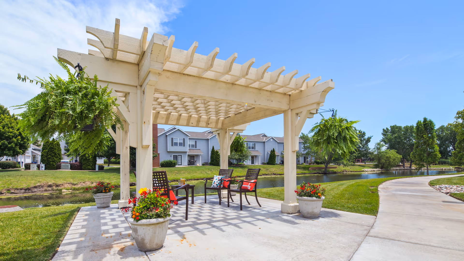 White wooden pergola with chairs, potted flowers, and hanging ferns beside a pond and walkway in a residential outdoor area.