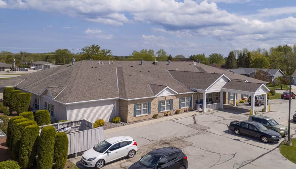 Exterior view of Harbor View Assisted Living facility showing a single-story building with a large roof, several windows with blue shutters, a covered entrance, and a parking lot with multiple cars. The surrounding area includes trimmed bushes, trees, and a partly cloudy sky.