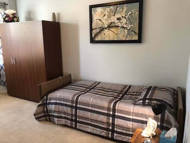Small bedroom with a single bed covered in a plaid comforter, a wooden wardrobe, framed artwork on the wall, and a nightstand with a lamp.