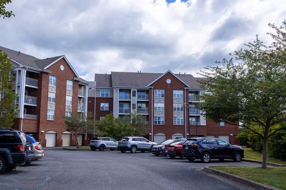 Exterior view of a multi-story brick residential building with balconies and multiple windows, surrounded by trees and a parking lot with several parked cars under a cloudy sky.