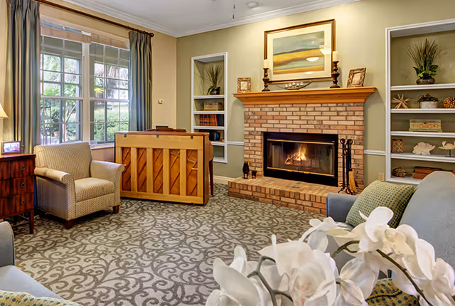 A cozy living room with a patterned carpet, a brick fireplace with a fire burning, built-in shelves decorated with plants and ornaments, a piano, an armchair, and a sofa. A large window with curtains lets in natural light.