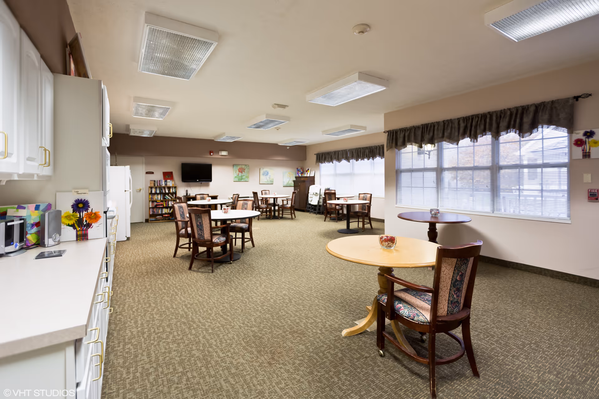 A spacious dining room with multiple round tables and chairs arranged throughout the room. Large windows with valance curtains allow natural light to fill the space. The room features a carpeted floor, white cabinetry along one wall with a microwave and flowers on the counter, and a television mounted on the far wall above a bookshelf. The ceiling has several rectangular fluorescent light fixtures.