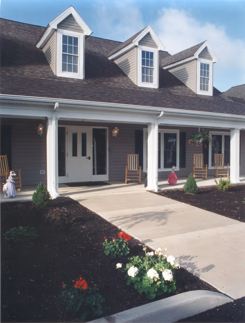 Covered front entrance of a residential building with a porch, rocking chairs, dormer windows, and flower beds lining the walkway.