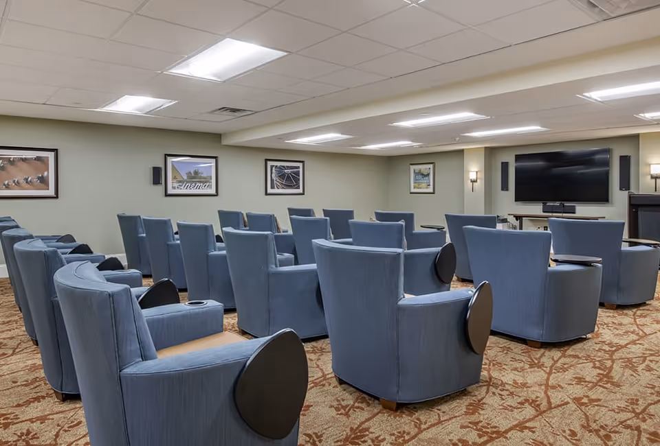 A small theater room with multiple rows of blue upholstered chairs facing a large flat-screen TV mounted on a beige wall. The room has a patterned carpet, framed pictures on the walls, and ceiling lights.