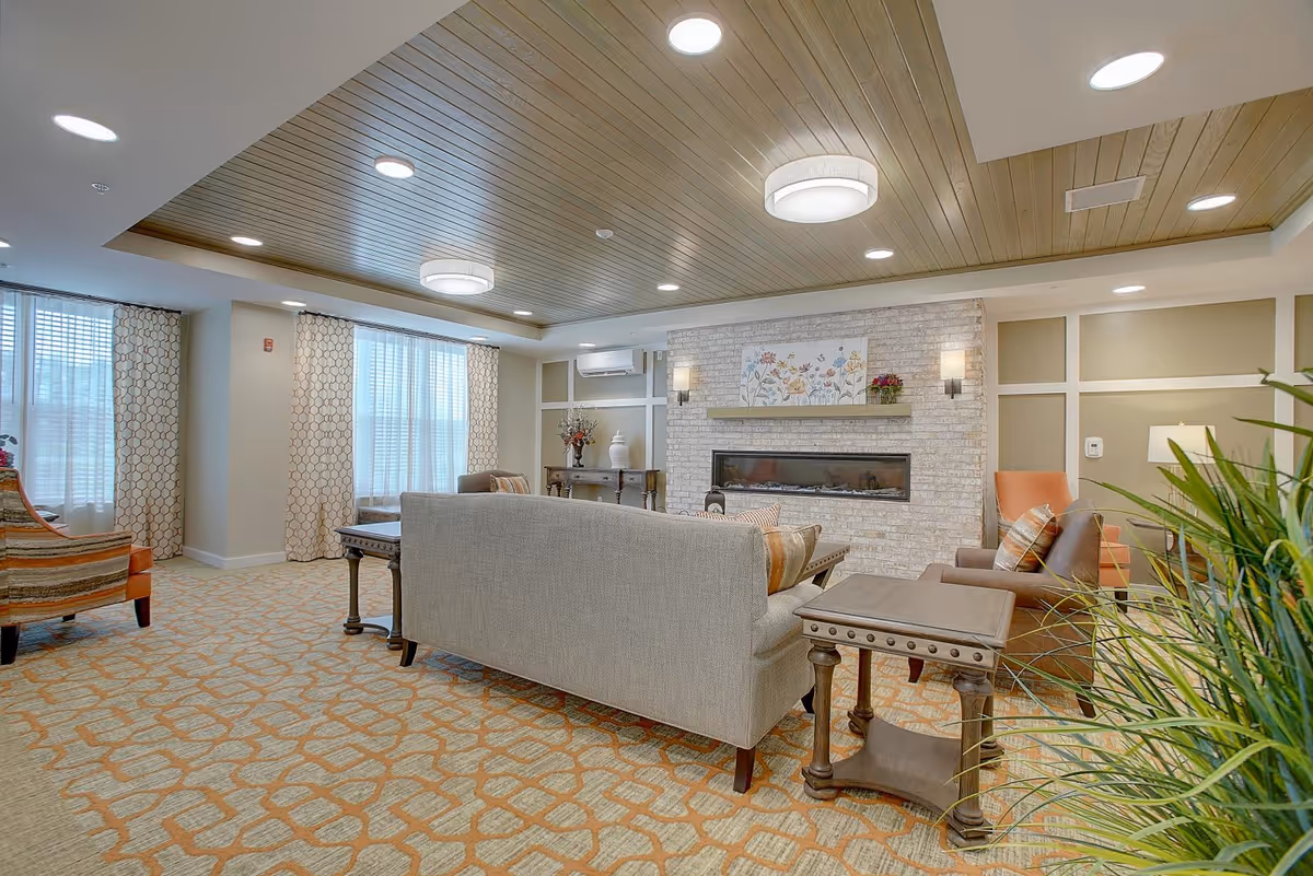 Bright communal living room with sofas and armchairs arranged around a long fireplace beneath a wood-paneled ceiling.