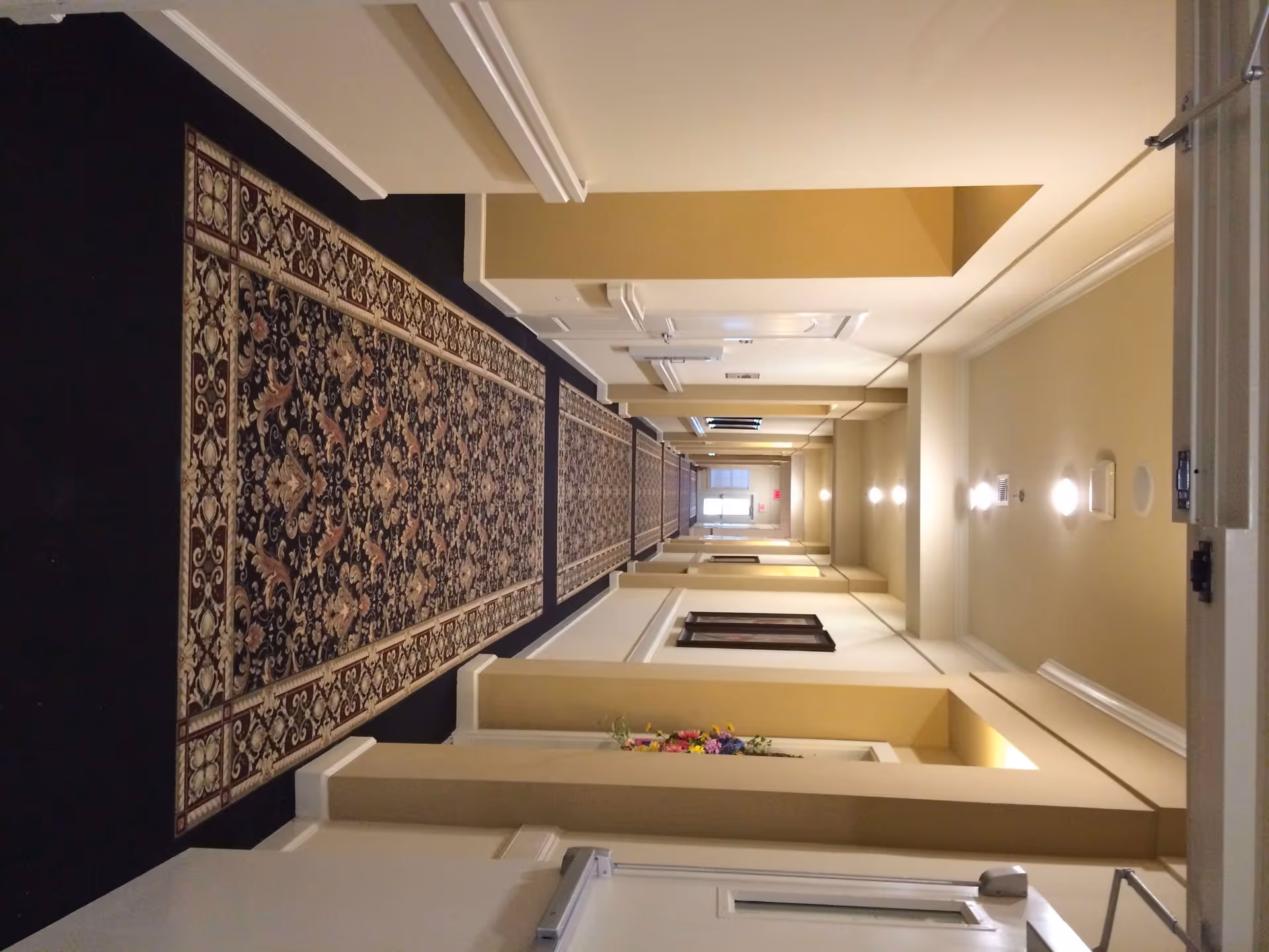A long, well-lit hallway with beige walls and a dark patterned carpet runner. The hallway has several doors on each side, framed artwork on the walls, and ceiling lights evenly spaced along the corridor.
