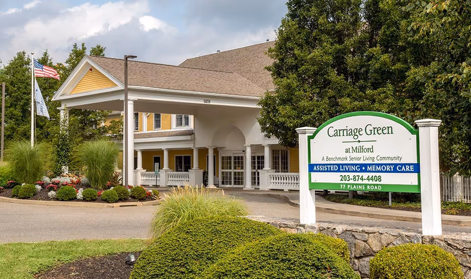 Exterior view of Carriage Green at Milford senior living community entrance with a covered driveway, landscaped bushes, and a sign displaying the facility name, services offered, phone number, and address.