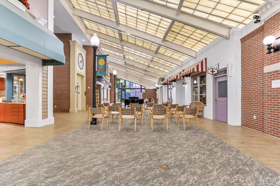 Interior view of a spacious common area in a senior living facility with a high slanted ceiling featuring large translucent panels. The area has a carpeted section with multiple wooden chairs arranged in rows, some with plaid cushions. On the left side, there is a kitchen or service counter with red cabinets and a teal awning. On the right side, there are storefront-style windows and doors, one with a red cross sign above it, suggesting a medical or nurse's station. The walls are a mix of white and red brick, and there are several lamppost-style lights along the corridor. A large clock is mounted on a brick wall near the center.