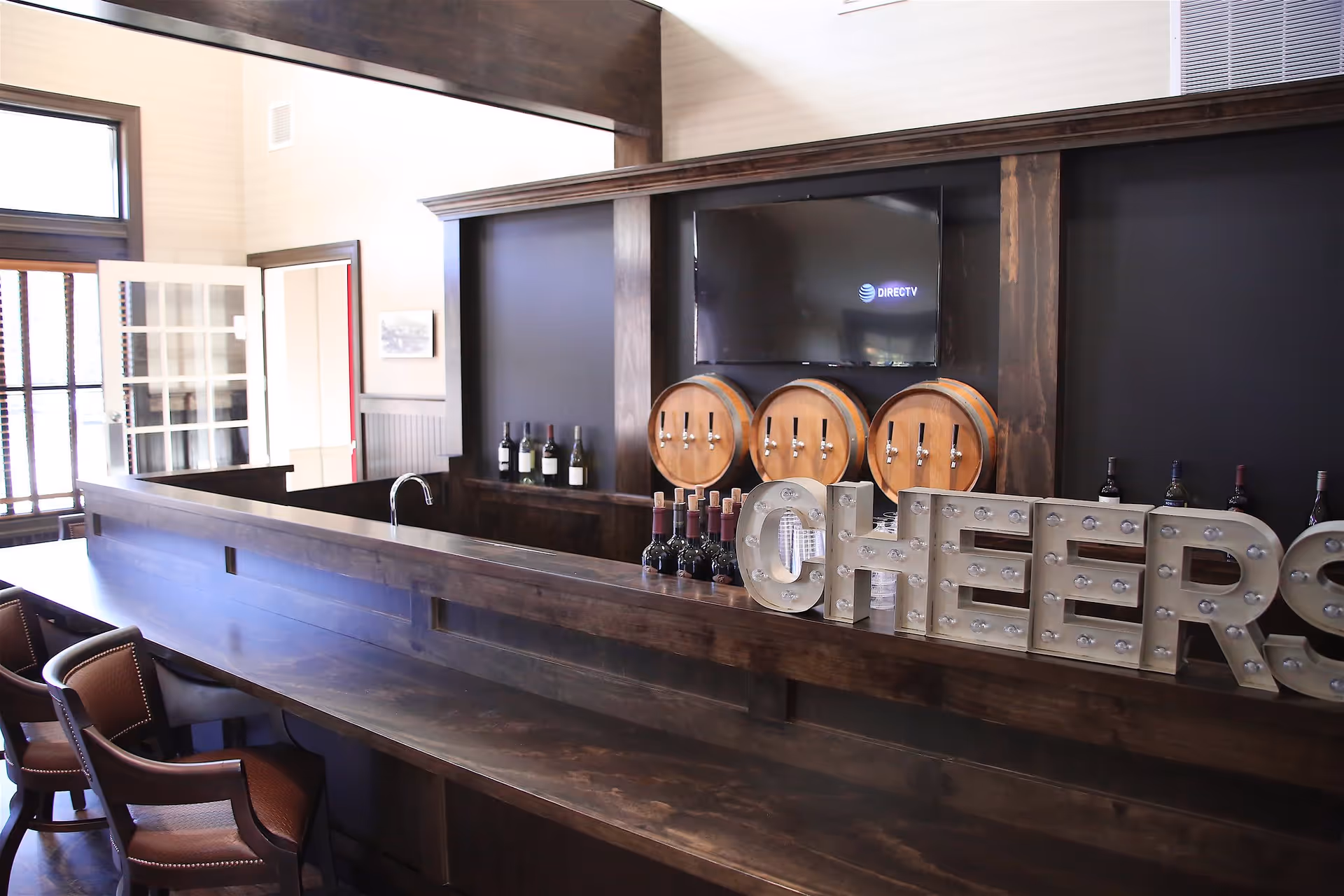 Wooden bar/lounge with chairs, wine bottles, barrel taps, a marquee 'CHEERS' sign and a wall-mounted TV.