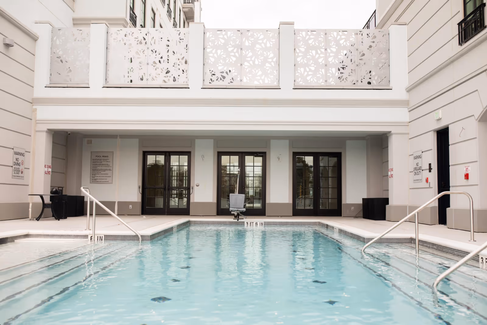 Outdoor swimming pool with clear blue water, surrounded by white walls and railings. There are three sets of double glass doors in the background, and decorative white panels above. Pool depth markers indicate 3 feet 6 inches. Safety signs are visible on the walls.
