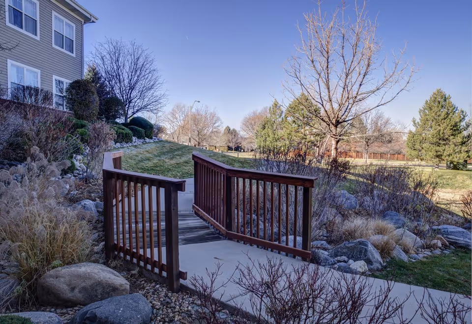 A small wooden footbridge over a landscaped garden area with rocks, bushes, and dry grasses. To the left is part of a multi-story building with beige siding and white-framed windows. In the background, there are leafless trees and evergreen trees under a clear blue sky.