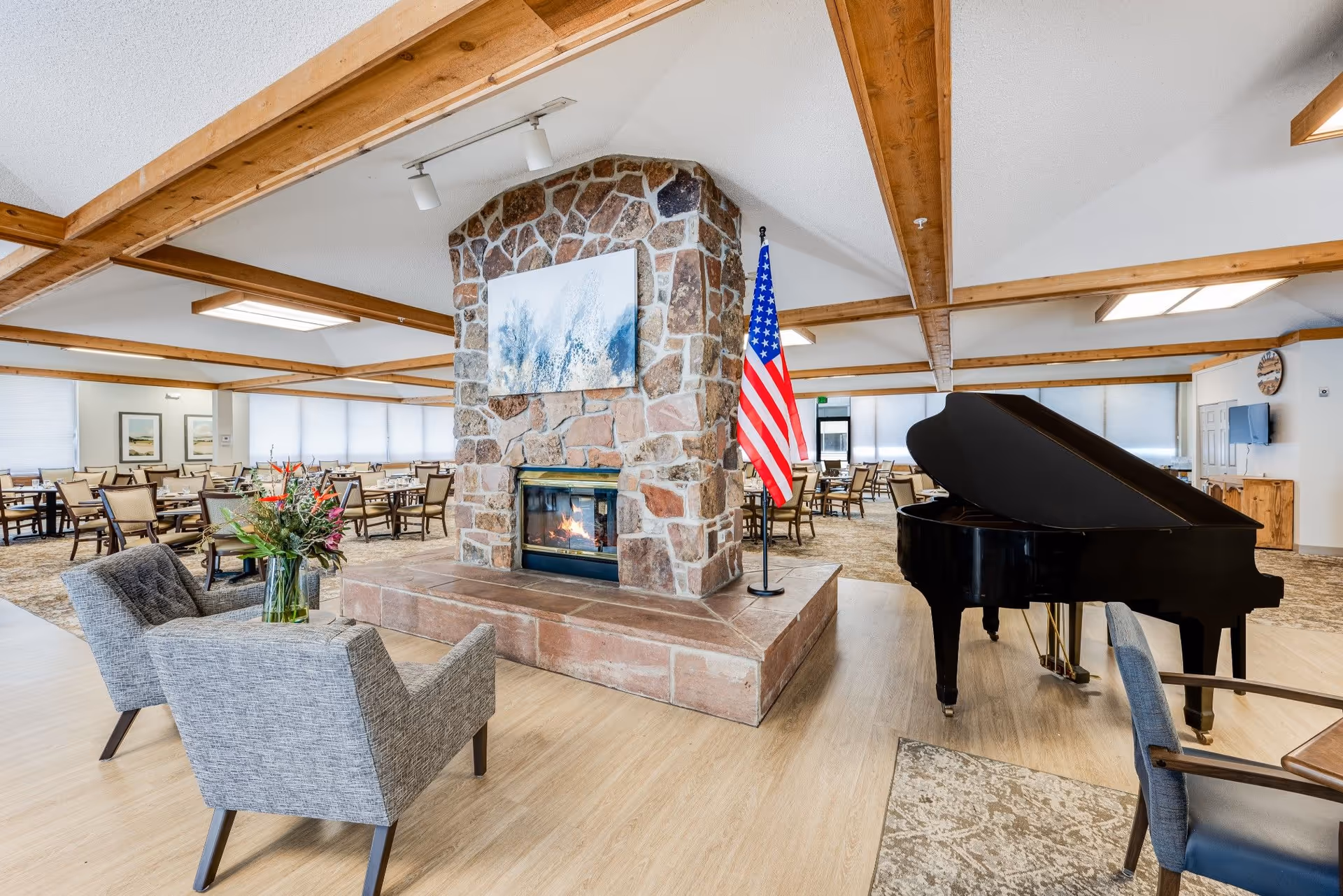 A spacious common area with a stone fireplace in the center, a burning fire, and an American flag beside it. There are two gray upholstered chairs with a vase of flowers on a small table between them in the foreground. To the right, there is a black grand piano. In the background, there are multiple dining tables and chairs arranged neatly, with large windows covered by blinds letting in natural light. The ceiling has wooden beams and modern light fixtures.