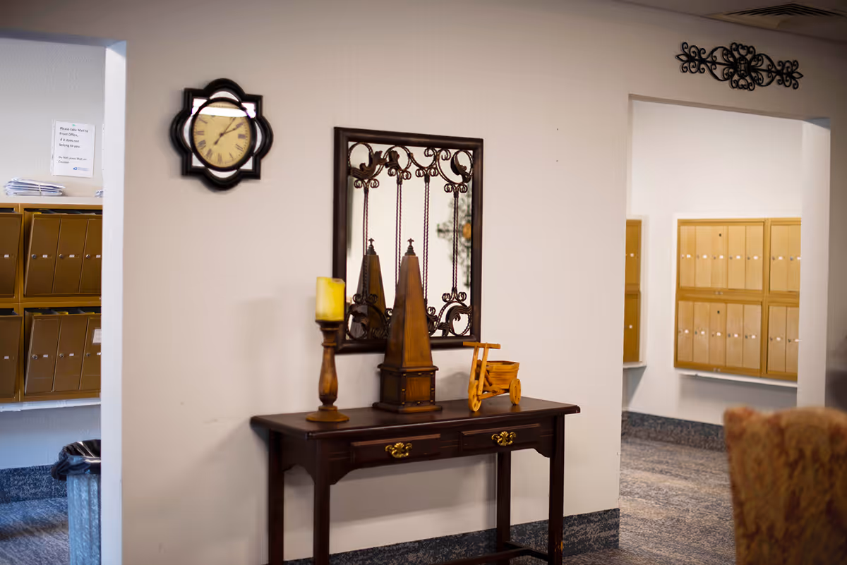 Interior hallway area with a wooden table against a white wall. On the table are decorative items including a candle holder, a wooden obelisk, and a small wooden tricycle. Above the table is a decorative mirror with an ornate metal frame. To the left and right of the table are mailboxes mounted on the walls. A clock with a decorative frame is mounted on the wall above the table. Part of an upholstered chair is visible in the lower right corner.