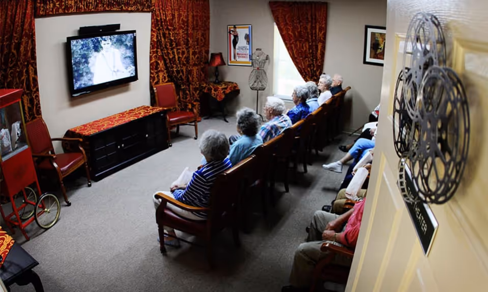 A group of elderly people seated in rows of chairs watching a black and white movie on a wall-mounted TV in a small theater room. The room has red and gold patterned curtains, a popcorn machine, and movie-themed decorations including film reels on the door.