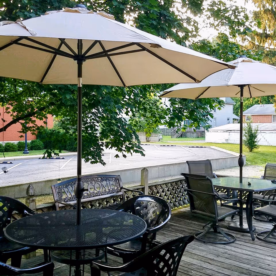 Outdoor patio area with black metal tables and chairs, each table shaded by a large beige umbrella. There is a wooden deck floor and a decorative bench with a 'WELCOME' sign. Trees and a parking lot with houses are visible in the background.