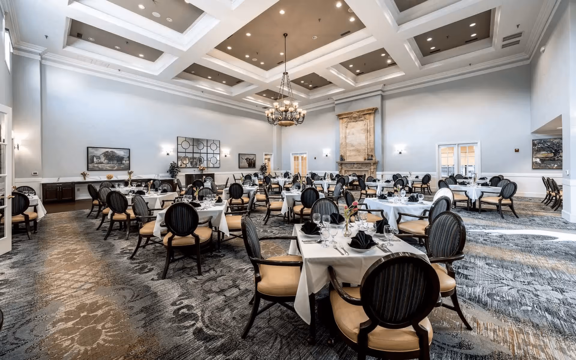 A spacious, elegant dining room with multiple round tables covered in white tablecloths, set with glassware, silverware, and black folded napkins. The room features high coffered ceilings with recessed lighting and a large chandelier. There is a decorative fireplace on one wall, framed artwork, and a patterned carpet covering the floor. The chairs have dark wooden frames with beige cushioned seats and striped backs.