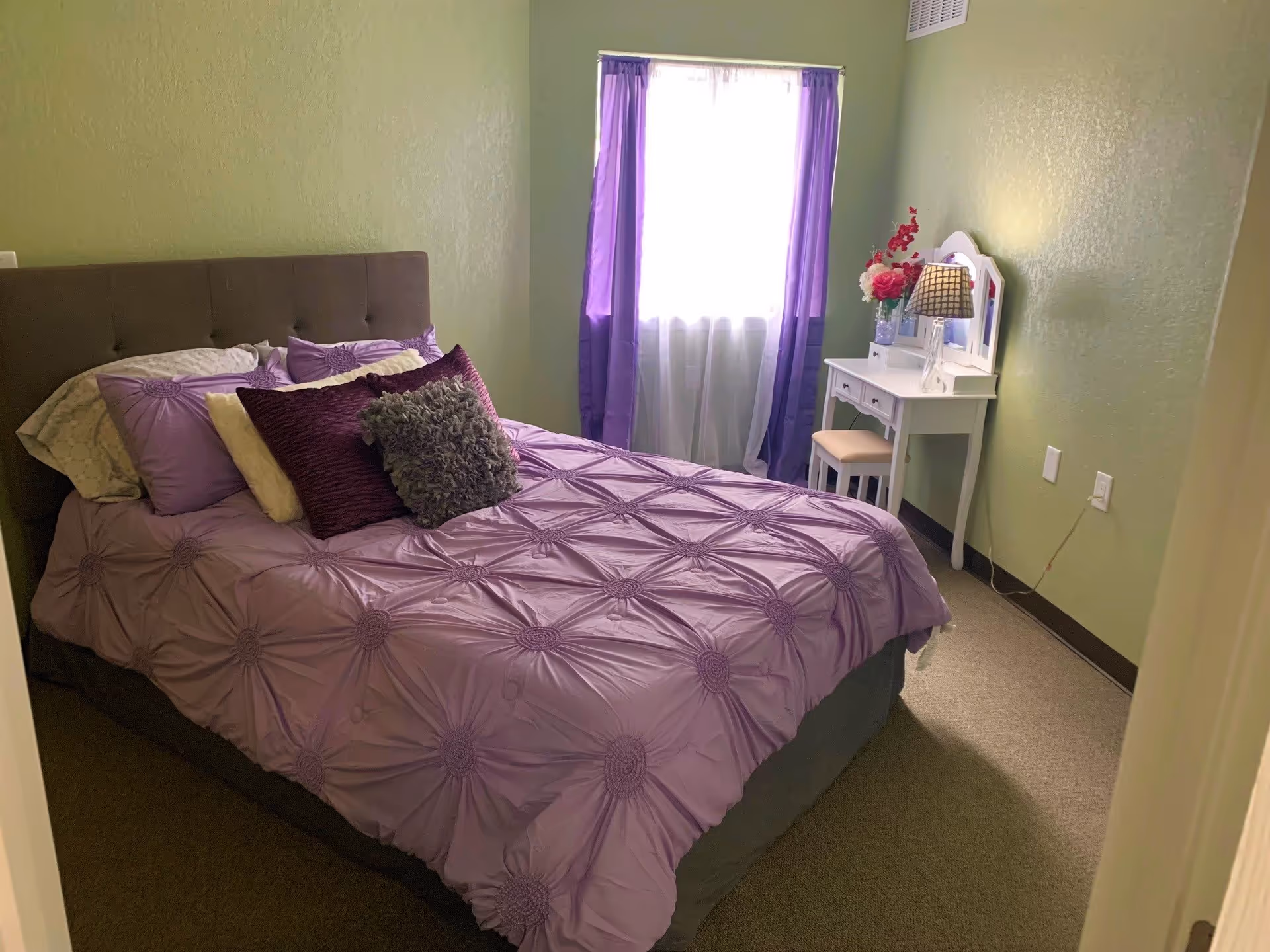 A bedroom with a bed covered in a purple comforter and multiple pillows. There is a window with purple curtains letting in natural light. A white vanity table with a mirror, a lamp, and a vase of flowers is positioned against the wall.