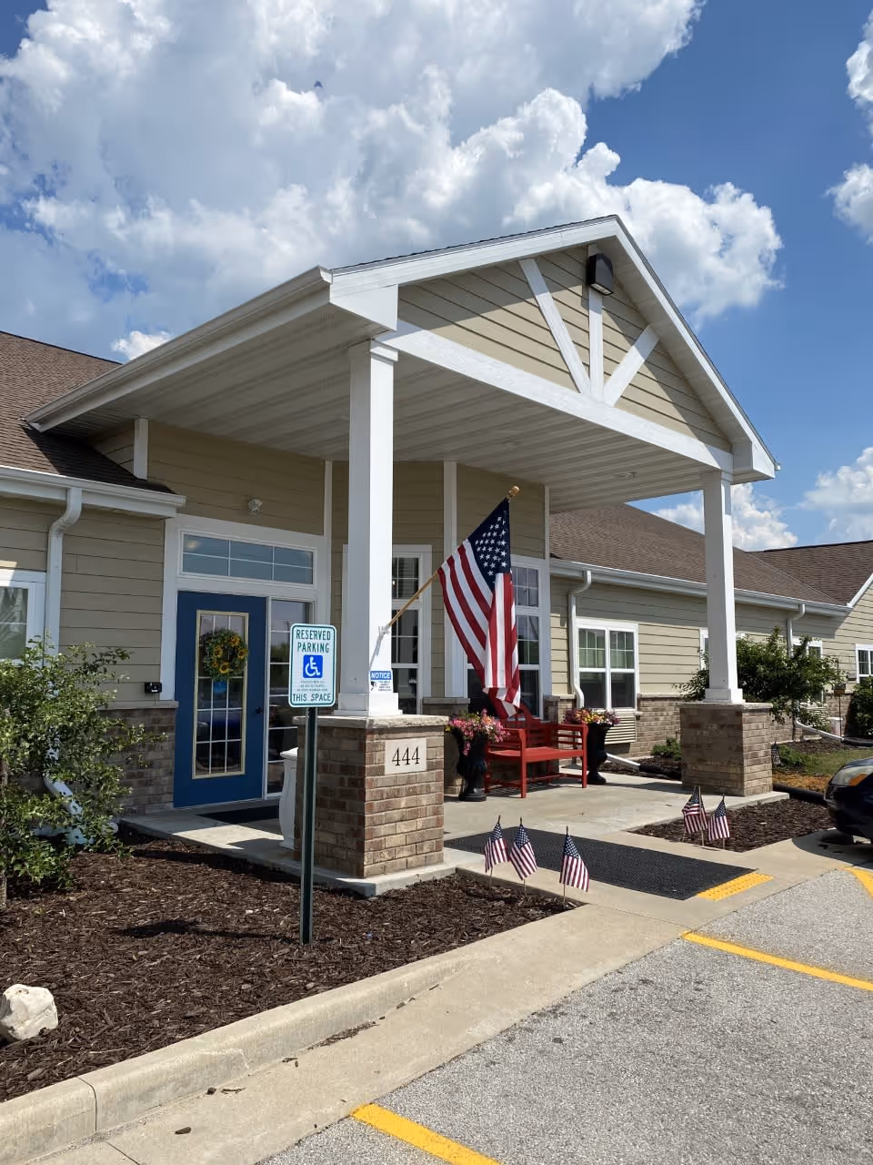 Front entrance of a single-story building with beige siding and brick pillars, featuring a covered porch with white columns. There is a blue door with a wreath, a red bench, and several American flags displayed around the entrance. A reserved parking sign for handicapped parking is visible near the sidewalk.
