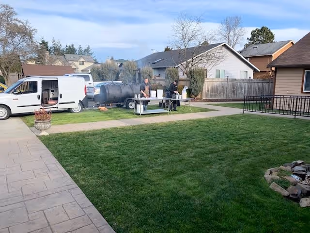 A grassy backyard area with a paved walkway and a fire pit made of stones. Two people are standing near a large barbecue smoker and a table with supplies. There are houses and a wooden fence in the background under a partly cloudy sky.