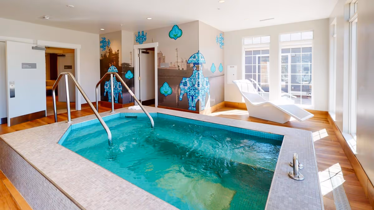 Indoor pool with clear turquoise water and metal handrails, surrounded by beige tiled edges and wooden flooring. The room has large windows allowing natural light to fill the space, a white lounge chair near the windows, and decorative blue wall art on the far wall.
