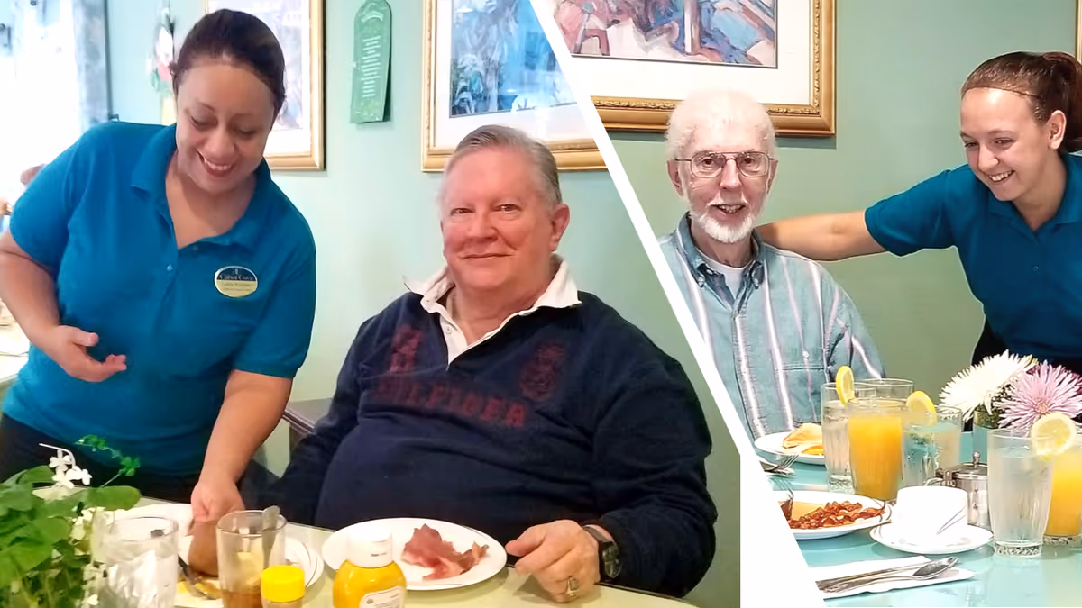 Two elderly men sitting at a dining table with plates of food and drinks, each accompanied by a smiling caregiver in teal uniforms. The setting appears to be a cheerful dining area with framed artwork on the walls and a vase of flowers on the table.