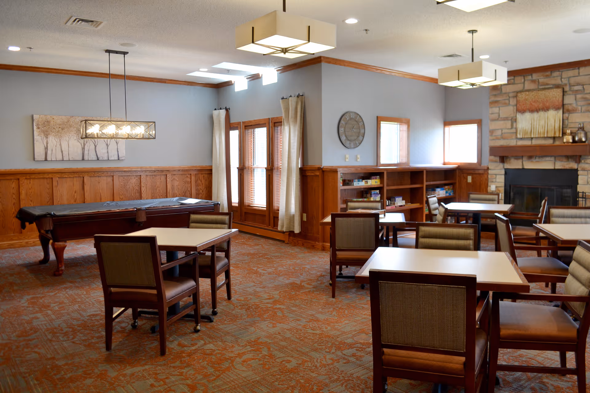 A spacious common room with several square tables and chairs arranged for group seating. The room features a pool table covered with a black cloth, a stone fireplace with a painting above it, wooden wainscoting on the walls, and large windows with curtains allowing natural light to enter. The carpet has a patterned design, and modern ceiling lights hang from the ceiling.