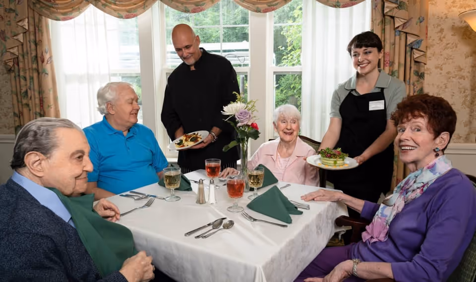 A group of senior residents seated around a table being served food by two staff members in a bright dining room.
