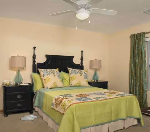 A bedroom with a black wooden headboard bed dressed in green bedding and floral pillows. There are two matching black nightstands on either side of the bed, each with a glass lamp. A ceiling fan with a light is mounted on the ceiling, and green curtains hang by a window on the right side of the room.