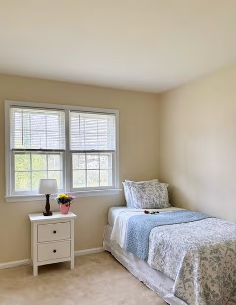 A small, neatly arranged bedroom with a single bed against the right wall. The bed is made with white and light blue floral bedding and a matching pillow. Next to the bed is a white nightstand with two drawers, holding a small table lamp and a pink pot with yellow flowers. Two large windows with white blinds allow natural light to fill the room. The walls are painted a light beige color and the floor is carpeted in a neutral tone.