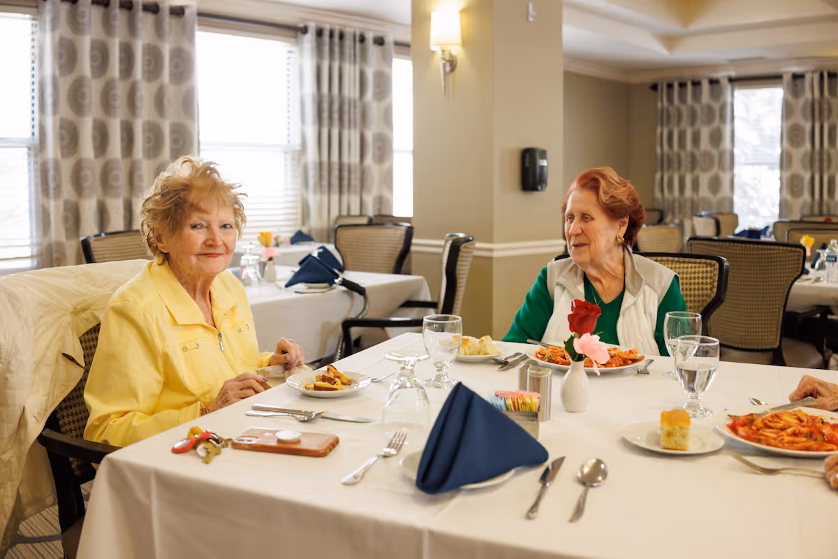 Two older women seated at a set table in a bright dining room, enjoying a meal.