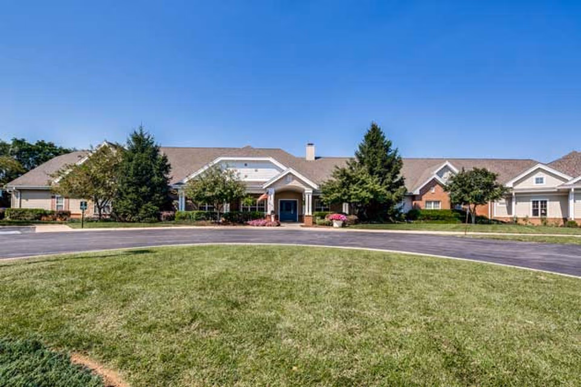 Single-story senior living facility building with a circular driveway, manicured lawn and trees under a clear blue sky.