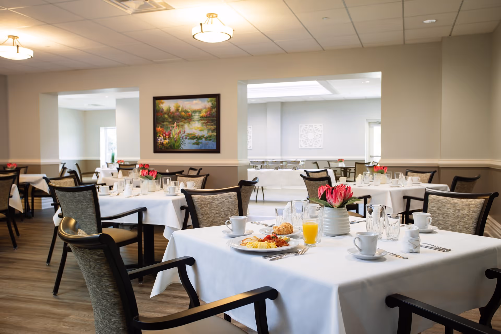 A bright dining room with multiple tables covered in white tablecloths, set with plates, cups, glasses, and silverware. Each table has a small vase with pink flowers. The room has light-colored walls, wooden flooring, and ceiling lights. A framed painting of a nature scene hangs on one wall.