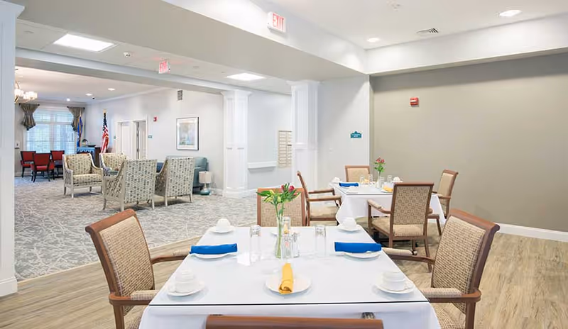 A bright and clean dining area in a senior living facility with tables set for meals, including white tablecloths, napkins, cups, and glasses. The room features comfortable wooden chairs with cushioned seats. In the background, there is a lounge area with patterned armchairs and an American flag near a window with curtains.
