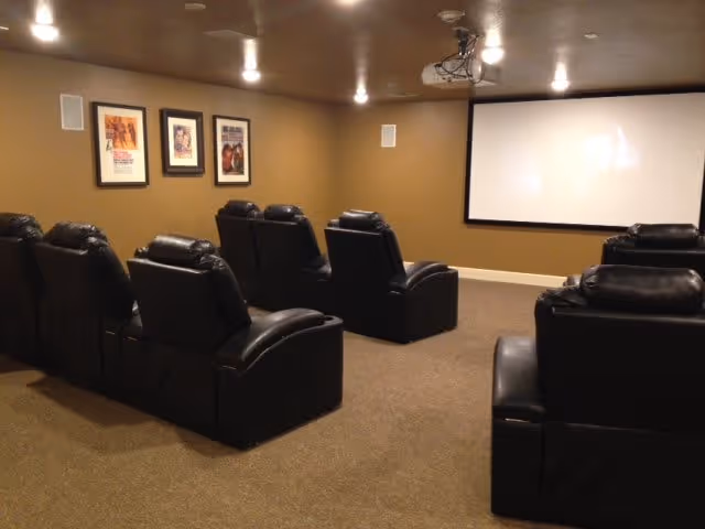 A small theater room with two rows of black leather recliner chairs facing a large white projection screen on a tan wall. The room has a brown carpet, tan walls, recessed ceiling lights, and three framed pictures on the left wall.