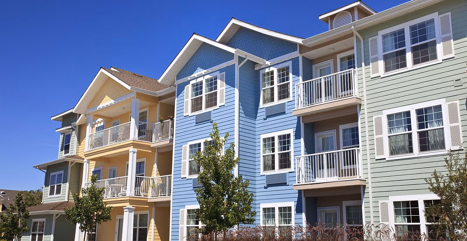 Three-story pastel-colored apartment building with balconies, windows, and trees under a clear blue sky.