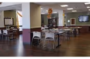 Spacious communal dining area with tables and chairs, wood flooring, shelving and a wall-mounted television.
