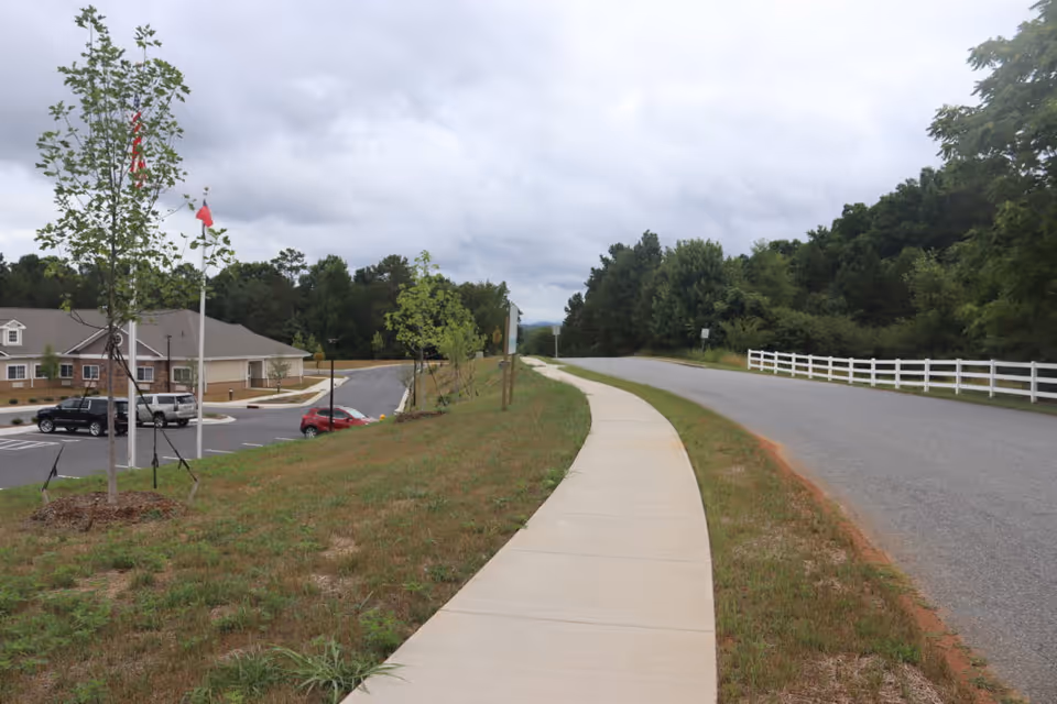 A curved sidewalk runs alongside a road past a parking lot and low building with flagpoles, trees, and a cloudy sky.