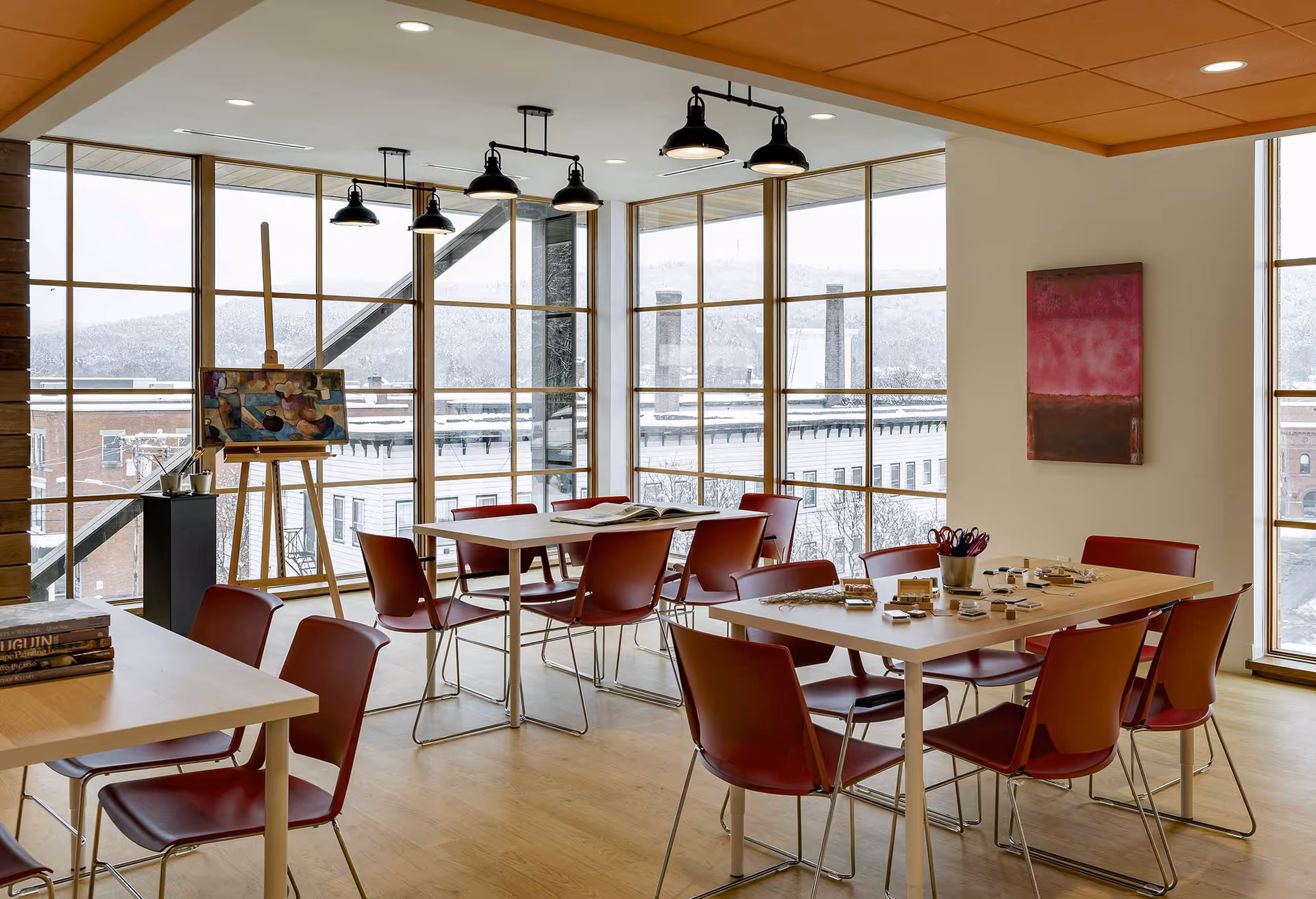 A bright room with large floor-to-ceiling windows showing a snowy outdoor view. Inside, there are several tables with red chairs arranged around them. One table has art supplies including paintbrushes and small containers, while another table has an open book. An easel with a colorful painting is positioned near the windows. The room has wooden floors, modern ceiling lights, and a pink and brown abstract painting on the wall.