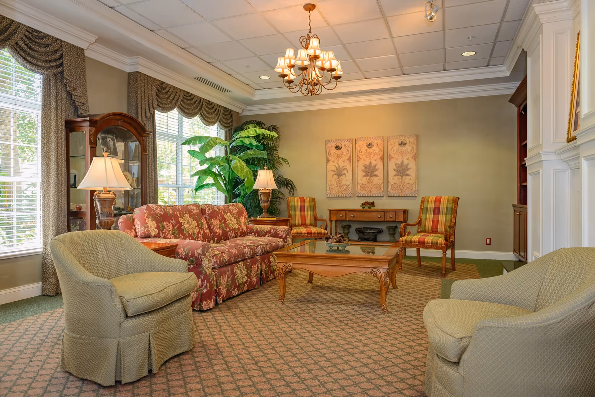 A cozy living room with a floral patterned sofa, two green armchairs, and two plaid chairs around a wooden coffee table with a glass top. The room has large windows with draped curtains, a wooden display cabinet, two table lamps, a chandelier, and decorative wall art above a wooden console table.