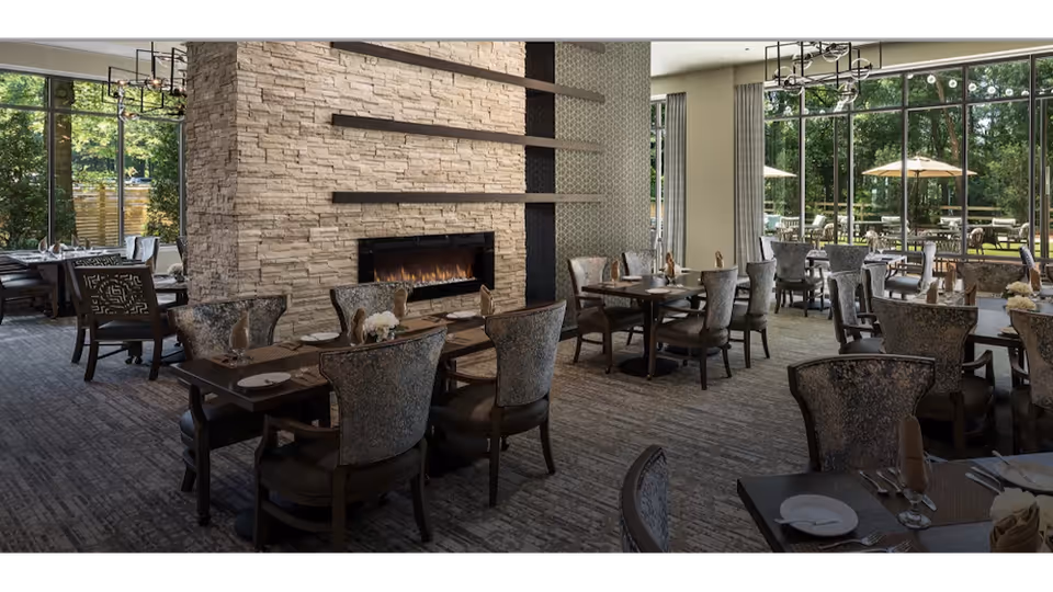 A dining room with multiple tables and chairs arranged neatly. The room features a large stone fireplace in the center, modern chandeliers hanging from the ceiling, and large floor-to-ceiling windows that provide a view of an outdoor patio with umbrellas and greenery.
