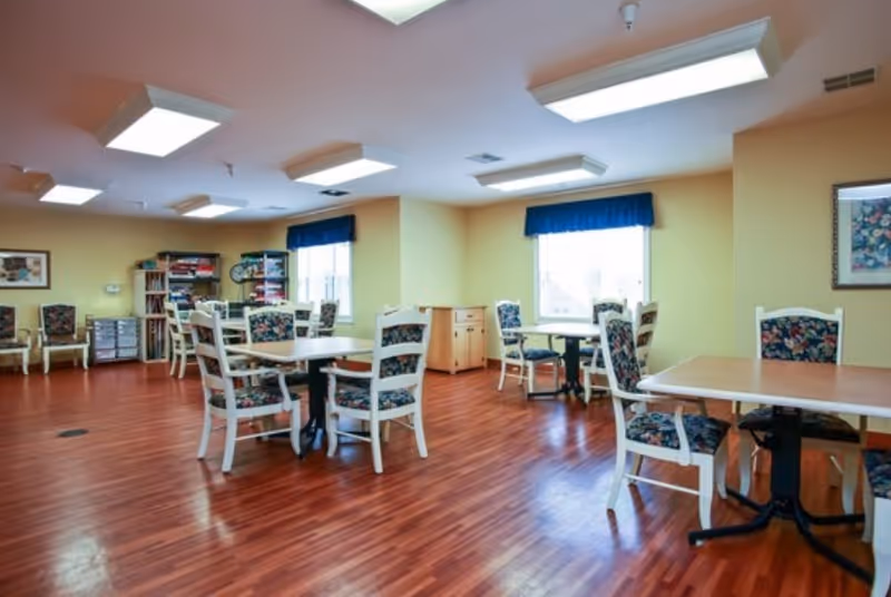 Bright communal dining/activity room with several tables and floral-upholstered chairs on polished wood floors under recessed lighting.