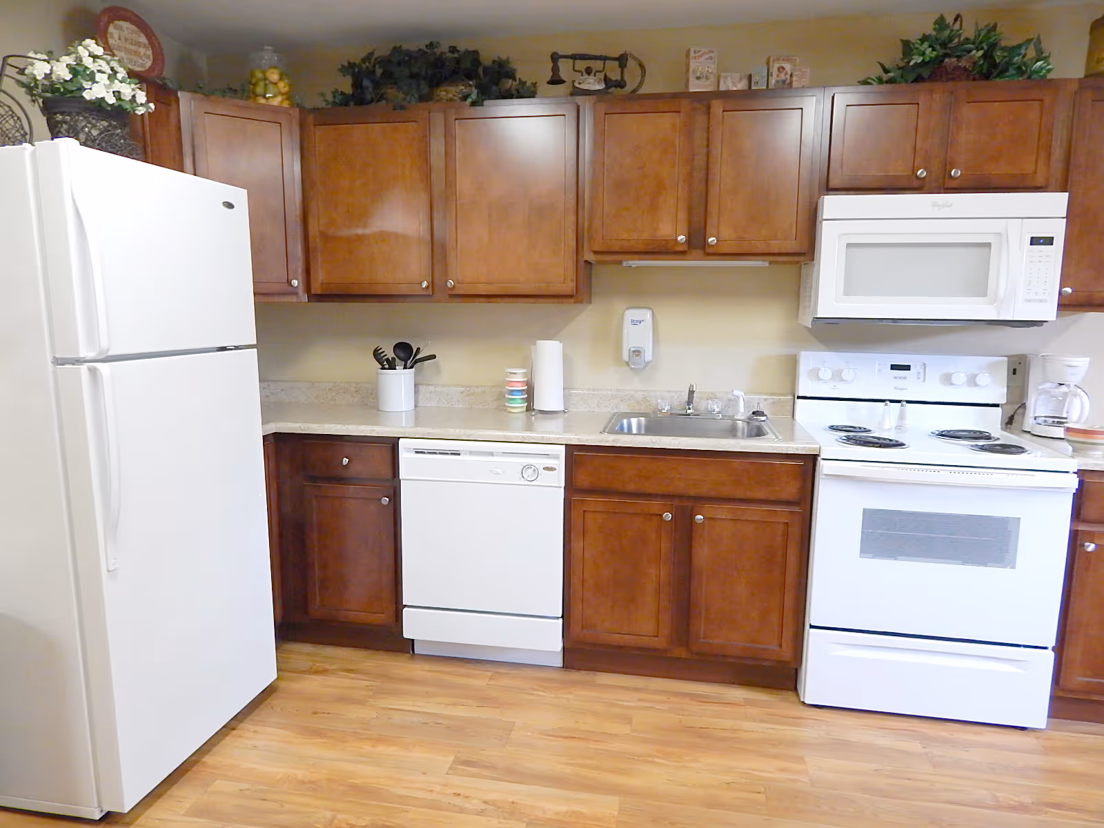 A kitchen with wooden cabinets, a white refrigerator, a white dishwasher, a white stove with an oven, a white microwave above the stove, a sink with a faucet, and various kitchen utensils and decorations on the countertop and above the cabinets.