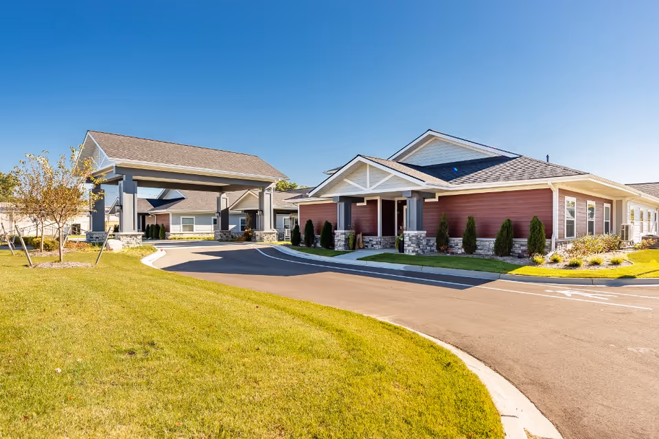 Exterior view of a senior living facility with a covered entrance driveway, manicured green lawn, small trees, and shrubs under a clear blue sky.