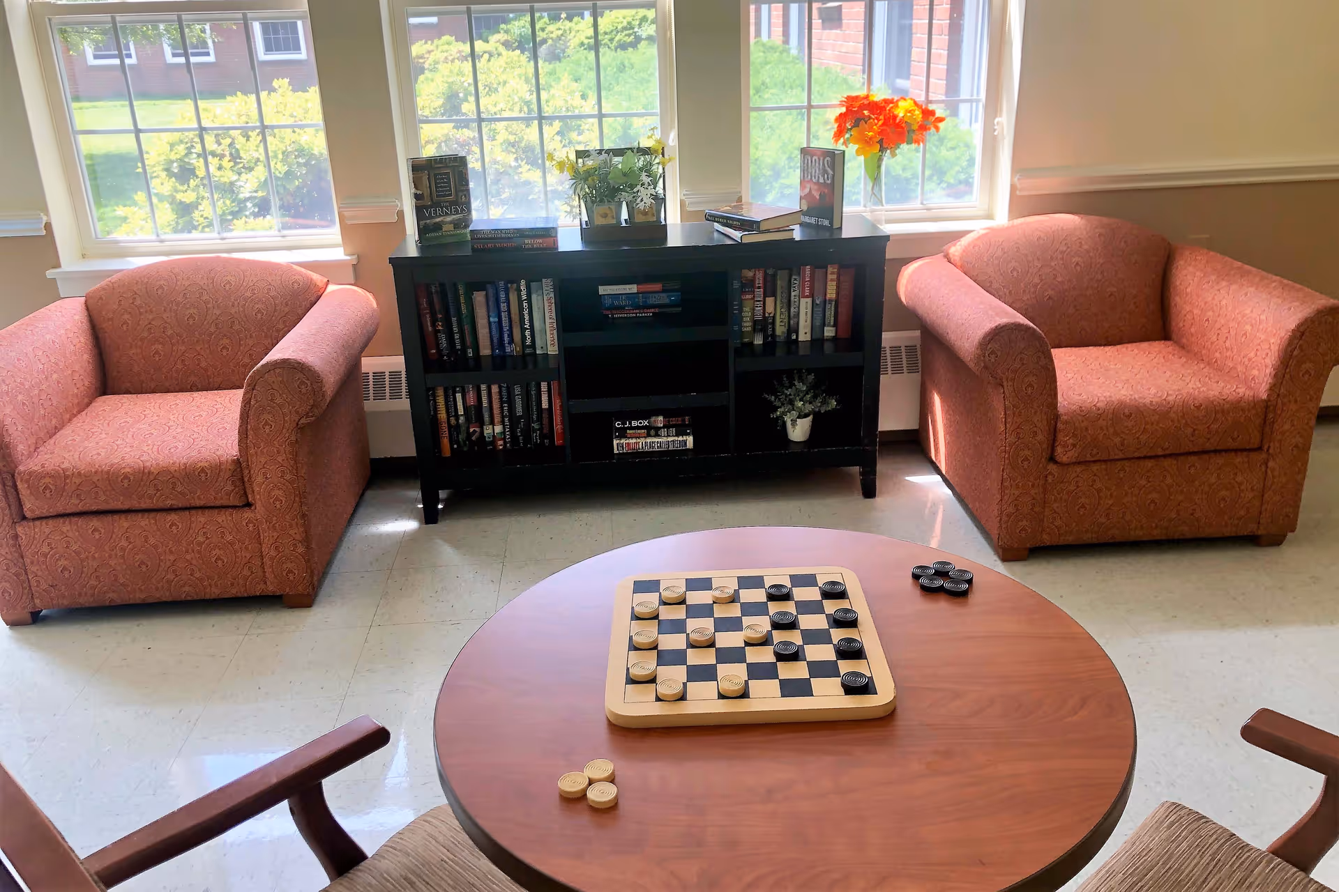 A cozy sitting area with two patterned armchairs facing a round wooden table with a checkers game set up. Behind the chairs is a black bookshelf filled with books and decorative plants, positioned in front of large windows letting in natural light and showing greenery outside.