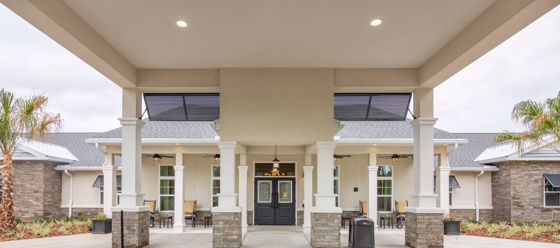 Entrance of The Addison of St. Johns facility showing a covered porch with white columns and stone bases, black double doors with decorative glass, outdoor seating with rocking chairs, ceiling fans, and palm trees on either side under a cloudy sky.