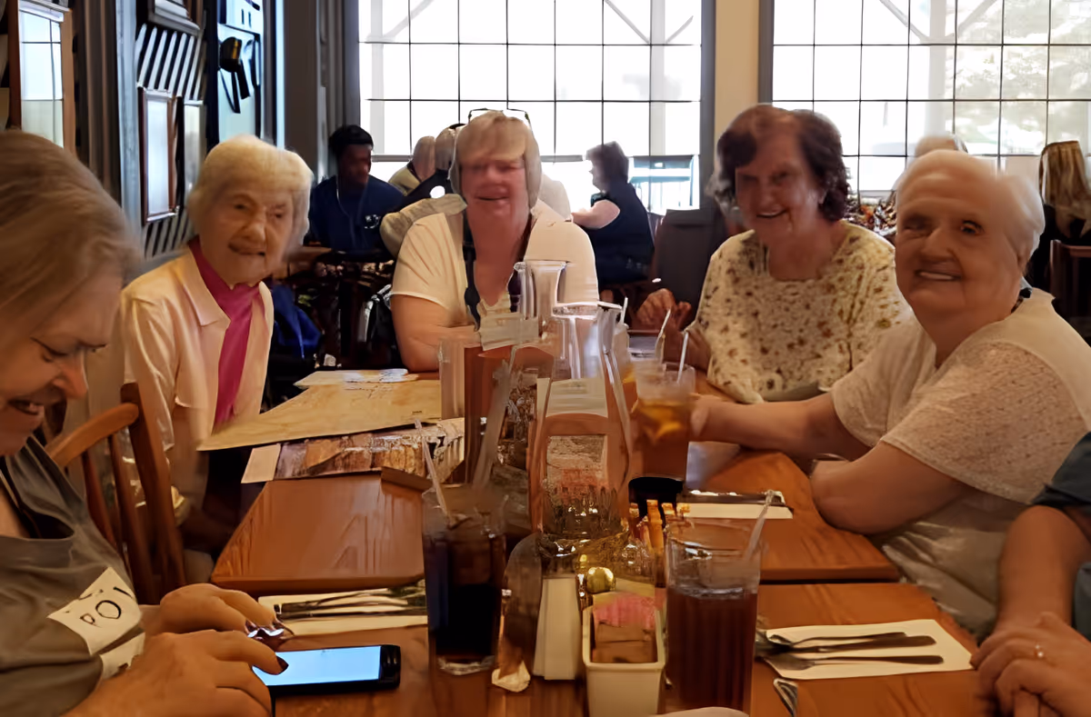A group of elderly women sitting around a wooden dining table in a well-lit room with large windows. They are smiling and appear to be enjoying each other's company. The table has several glasses of iced tea, menus, and condiments.