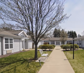 A paved walkway through a courtyard with trimmed bushes and leafless trees, surrounded by single-story white buildings with multiple windows under a clear sky. A white metal bench is placed near the walkway.