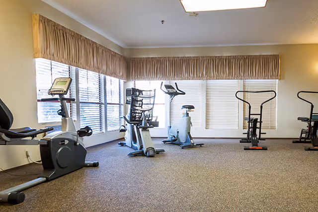 A small fitness room with several exercise bikes positioned on a carpeted floor. Large windows with blinds and beige valances allow natural light to enter the room.