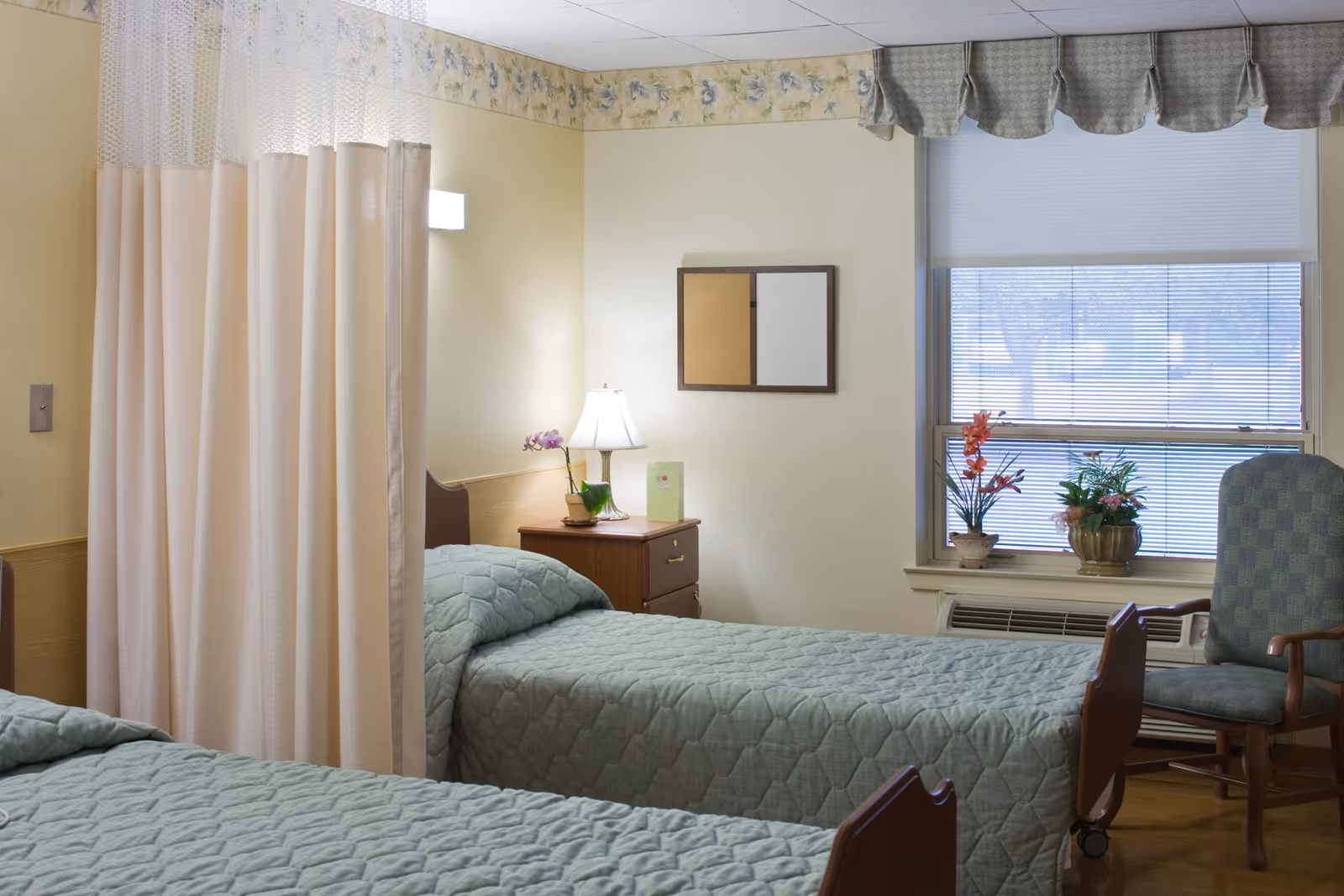 Two twin beds in a softly decorated senior living bedroom with a privacy curtain, nightstand and a window with plants.